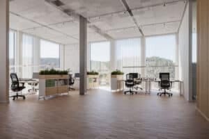 A spacious modern open-plan office with large floor-to-ceiling windows overlooking forested hills. Work desks with black task chairs are separated by low cabinets featuring wood and pale green accents, topped with planters full of greenery. The ceiling is a perforated acoustic panel design.